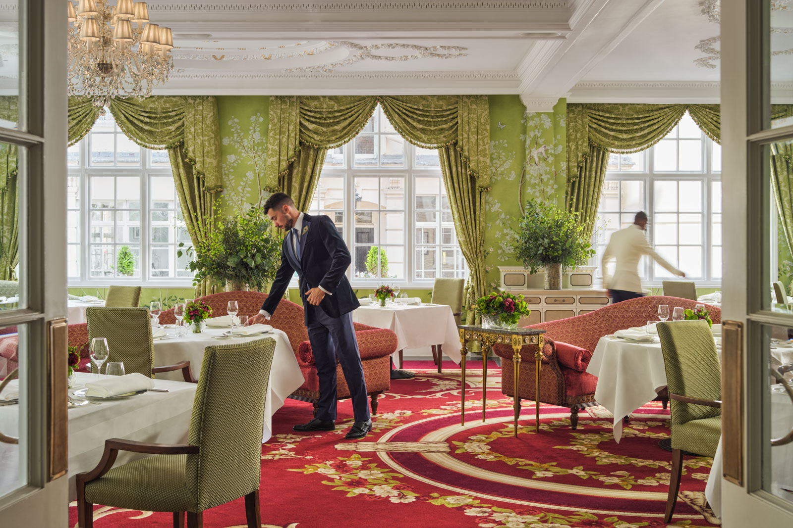 A man in a suit straightens objects on a table in a carpeted restaurant dining room.